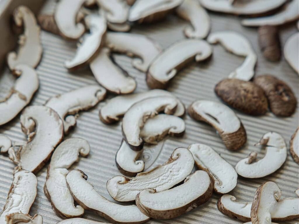 Sliced mushrooms are spread out in a single layer on a ridged, light-colored baking sheet, ready for roasting or drying. The mushrooms appear fresh and uncooked.